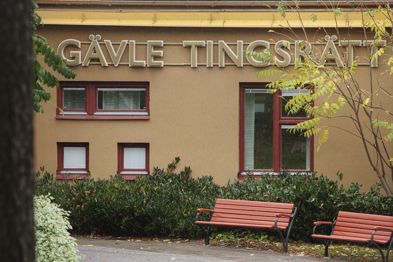 Gävle tingsrätts fasad med gula bokstäver och fönster omgivna av gröna buskar och parkbänkar i förgrunden.